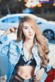 A woman in a black bra top and denim jacket posing next to a white car.